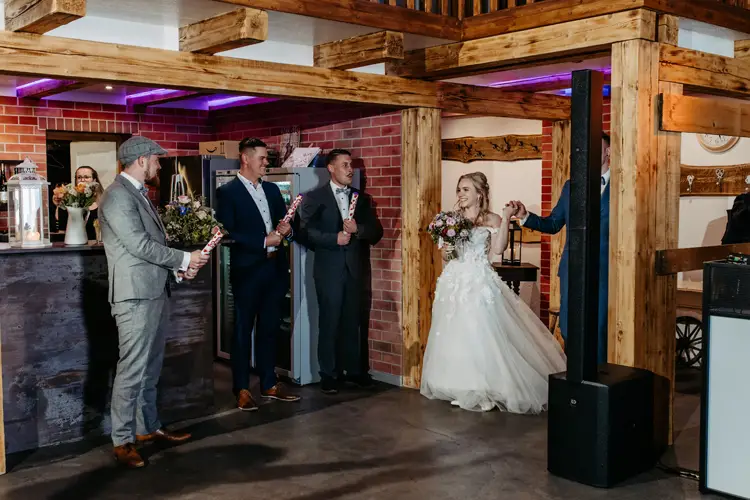 Eine Braut im weißen Kleid und ein Bräutigam im blauen Anzug tanzen im Innenbereich, während Brautjungfern in der Nähe stehen und Schilder mit der Aufschrift „Herr & Frau“ und „Herzlichen Glückwunsch“ hochhalten. Die Atmosphäre ist rustikal und mit Holzbalken geschmückt.