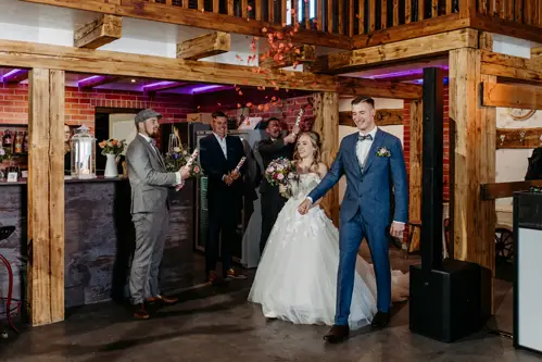 Eine Braut im weißen Kleid und ein Bräutigam im blauen Anzug tanzen im Innenbereich, während Brautjungfern in der Nähe stehen und Schilder mit der Aufschrift „Herr & Frau“ und „Herzlichen Glückwunsch“ hochhalten. Die Atmosphäre ist rustikal und mit Holzbalken geschmückt.