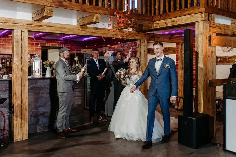 Eine Braut im weißen Kleid und ein Bräutigam im blauen Anzug tanzen im Innenbereich, während Brautjungfern in der Nähe stehen und Schilder mit der Aufschrift „Herr & Frau“ und „Herzlichen Glückwunsch“ hochhalten. Die Atmosphäre ist rustikal und mit Holzbalken geschmückt.
