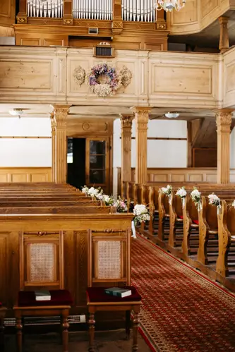 Ein hölzerner Kircheninnenraum mit mit kleinen Blumensträußen geschmückten Kirchenbankreihen, einem rot gemusterten Gangläufer und einer Orgel über dem Eingang im hinteren Bereich.