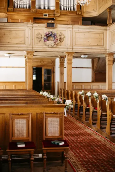 Ein hölzerner Kircheninnenraum mit mit kleinen Blumensträußen geschmückten Kirchenbankreihen, einem rot gemusterten Gangläufer und einer Orgel über dem Eingang im hinteren Bereich.