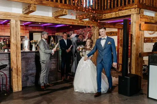 Eine Braut im weißen Kleid und ein Bräutigam im blauen Anzug tanzen im Innenbereich, während Brautjungfern in der Nähe stehen und Schilder mit der Aufschrift „Herr & Frau“ und „Herzlichen Glückwunsch“ hochhalten. Die Atmosphäre ist rustikal und mit Holzbalken geschmückt.