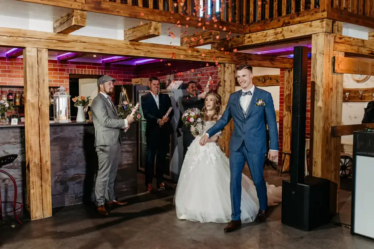 Eine Braut im weißen Kleid und ein Bräutigam im blauen Anzug tanzen im Innenbereich, während Brautjungfern in der Nähe stehen und Schilder mit der Aufschrift „Herr & Frau“ und „Herzlichen Glückwunsch“ hochhalten. Die Atmosphäre ist rustikal und mit Holzbalken geschmückt.