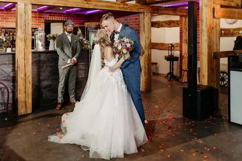 Eine Braut und ein Bräutigam küssen sich auf der Tanzfläche ihrer Hochzeit, umgeben von Holzbalken und Rosenblättern, während ein Mann in Anzug und Hut im Hintergrund steht und ihnen zusieht.