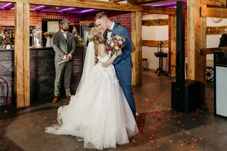 Eine Braut und ein Bräutigam küssen sich auf der Tanzfläche ihrer Hochzeit, umgeben von Holzbalken und Rosenblättern, während ein Mann in Anzug und Hut im Hintergrund steht und ihnen zusieht.