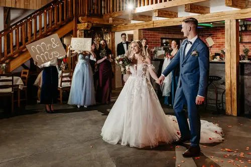 Eine Braut im weißen Kleid und ein Bräutigam im blauen Anzug tanzen im Innenbereich, während Brautjungfern in der Nähe stehen und Schilder mit der Aufschrift „Herr & Frau“ und „Herzlichen Glückwunsch“ hochhalten. Die Atmosphäre ist rustikal und mit Holzbalken geschmückt.