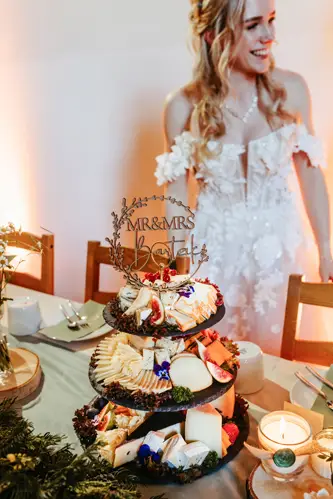Eine lächelnde Braut in einem weißen Spitzenkleid steht neben einer dreistöckigen Käseplatte mit einem „Mr & Mrs“-Schild, umgeben von Kerzen, Grünpflanzen und einem dekorierten Hochzeitstisch.