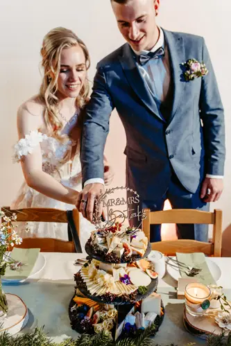 Lächelnd schneiden Braut und Bräutigam eine mehrschichtige Hochzeitstorte an, die mit Früchten und Käse dekoriert ist. Die Braut trägt ein schulterfreies Kleid, der Bräutigam einen blauen Anzug. Der Tisch ist mit grünen Servietten und Blumenarrangements gedeckt.