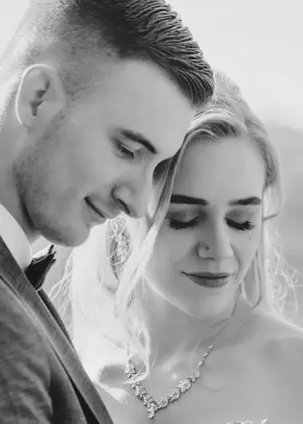 Eine Schwarz-Weiß-Nahaufnahme von Braut und Bräutigam, die eng beieinander stehen, beide sanft lächeln und ihre Augen niedergeschlagen haben. So wird ein intimer und zärtlicher Hochzeitsmoment eingefangen.