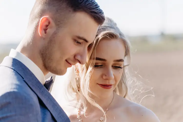 Eine Nahaufnahme von Braut und Bräutigam im Freien. Sie stehen eng beieinander und blicken mit sanftem Gesichtsausdruck nach unten. Der Bräutigam trägt einen hellgrauen Anzug, die Braut hat langes blondes Haar und eine juwelenbesetzte Halskette.