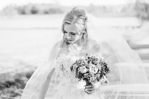Eine Braut in weißem Brautkleid und Schleier blickt mit einem Blumenstrauß in der Hand nach unten und steht im hellen Sonnenlicht im Freien. Das Bild ist schwarz-weiß.