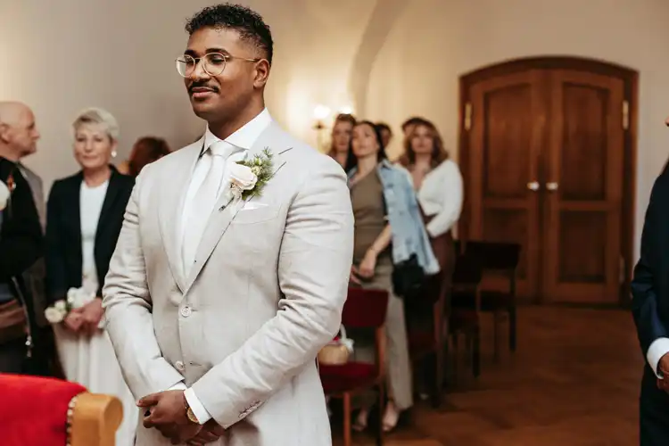 Ein Bräutigam in hellem Anzug und weißer Krawatte steht lächelnd vor dem Altar bei der Hochzeit in Görlitz. Hinter ihm versammeln sich die Gäste in einer warm beleuchteten Indoor-Zeremonie, perfekt eingefangen von Hochzeitsfotograf Görlitz.