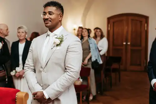 Ein Bräutigam in hellem Anzug und weißer Krawatte steht lächelnd vor dem Altar bei der Hochzeit in Görlitz. Hinter ihm versammeln sich die Gäste in einer warm beleuchteten Indoor-Zeremonie, perfekt eingefangen von Hochzeitsfotograf Görlitz.