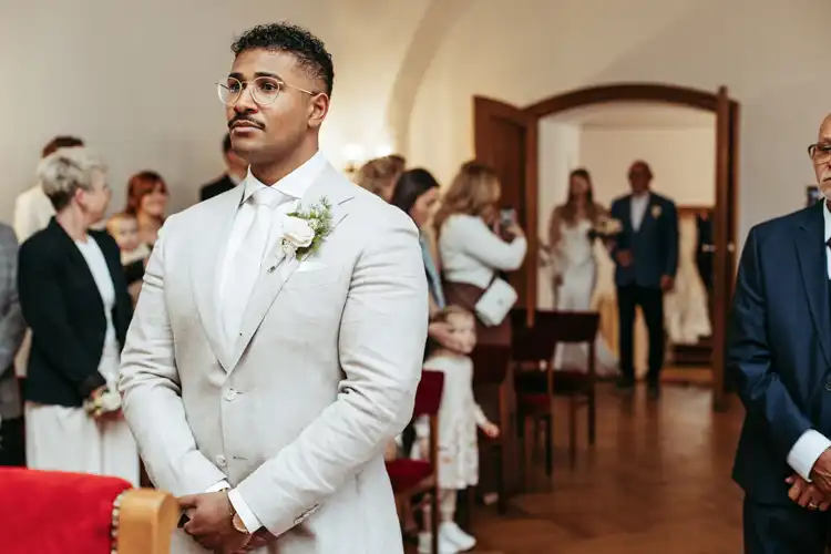 Ein Bräutigam im hellen Anzug steht vor dem Altar und blickt erwartungsvoll nach vorne. Die Hochzeitsgäste beobachten aus dem warm beleuchteten Raum, wie sich der besondere Tag auf Hochzeit in Görlitz entfaltet, festgehalten von einem talentierten Hochzeitsfotografen Görlitz.