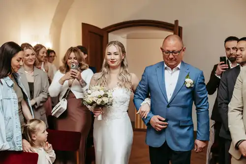 Auf der Hochzeit in Görlitz schreitet eine Braut in einem weißen Kleid mit einem Blumenstrauß in der Hand Arm in Arm mit einem Mann in einem blauen Anzug den Gang entlang. Die Gäste schauen zu und machen Fotos, während ein kleines Mädchen in der ersten Reihe bewundernd nach oben schaut - eingefangen von Hochzeitsfotograf Görlitz.