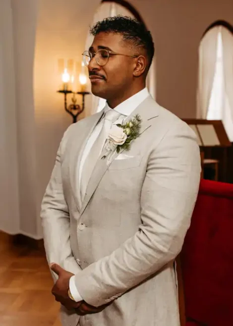 Ein Mann in einem hellbeigen Anzug, weißem Hemd und Krawatte steht mit gefalteten Händen in einem Haus. Er trägt eine Brille und eine Boutonnière und blickt ruhig vor sich hin. Elegante Beleuchtung und gewölbte Fenster bilden den Rahmen für eine Hochzeit in Görlitz, aufgenommen von Hochzeitsfotograf Görlitz.