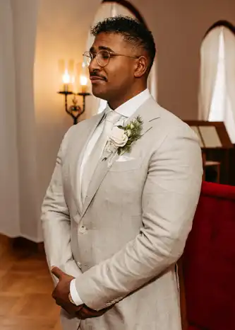 Ein Mann in einem hellbeigen Anzug, weißem Hemd und Krawatte steht mit gefalteten Händen in einem Haus. Er trägt eine Brille und eine Boutonnière und blickt ruhig vor sich hin. Elegante Beleuchtung und gewölbte Fenster bilden den Rahmen für eine Hochzeit in Görlitz, aufgenommen von Hochzeitsfotograf Görlitz.