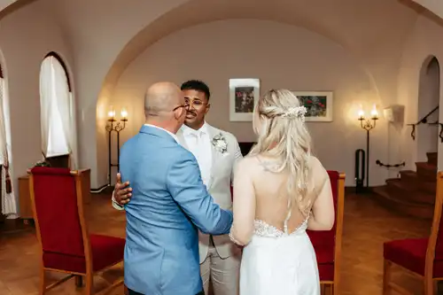 Ein Bräutigam im weißen Anzug umarmt einen älteren Mann in einer blauen Jacke, während die Braut mit langen blonden Haaren neben ihnen in einem eleganten Raum mit roten Stühlen und Bogenfenstern auf ihrer Hochzeit in Görlitz steht. Hochzeitsfotograf Görlitz hält den Moment fest.