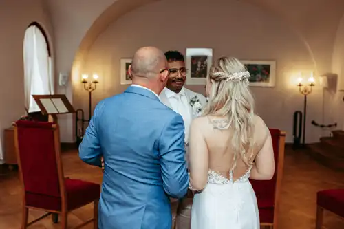 Eine Braut in einem weißen Kleid steht mit einem Mann in einem blauen Anzug vor einem lächelnden Bräutigam in einem hellen Anzug während einer Hochzeitszeremonie in Görlitz, wunderschön fotografiert von Hochzeitsfotograf Görlitz.