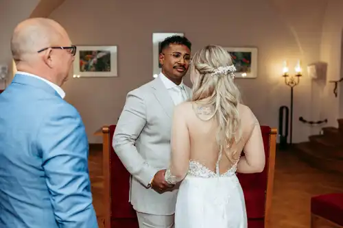 Ein Bräutigam in einem hellen Anzug lächelt und hält die Hand seiner Braut, die ein weißes Kleid mit offenem Rücken trägt. In der Nähe steht ein Mann in einem blauen Anzug in dem warm beleuchteten Raum von Hochzeit Gut am See, gerahmte Kunst an den Wänden. Hochzeitsfotograf Görlitz hält den Moment fest.