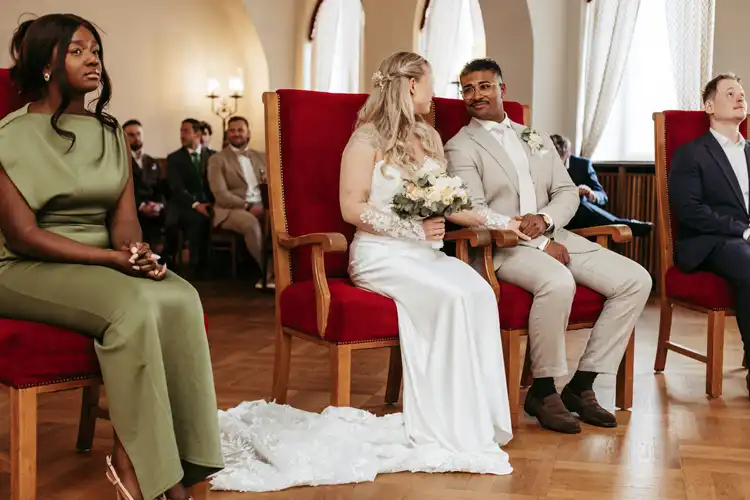Eine Braut und ein Bräutigam sitzen während einer Hochzeitszeremonie im Standesamt Görlitz auf roten Stühlen, halten sich an den Händen und lächeln. Eine Frau in einem grünen Kleid sitzt links von ihnen, ein Mann im Anzug rechts von ihnen. Die Gäste sitzen im Hintergrund.