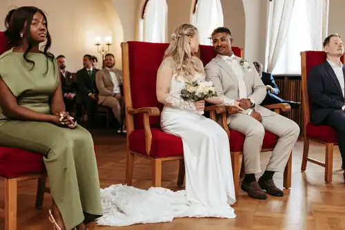 Eine Braut und ein Bräutigam sitzen während einer Hochzeitszeremonie im Standesamt Görlitz auf roten Stühlen, halten sich an den Händen und lächeln. Eine Frau in einem grünen Kleid sitzt links von ihnen, ein Mann im Anzug rechts von ihnen. Die Gäste sitzen im Hintergrund.