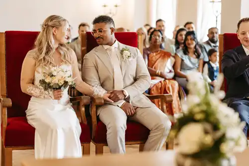 Eine Braut und ein Bräutigam sitzen händchenhaltend und lächeln sich an, während sie ihre Hochzeit in Görlitz feiern, während die Gäste im Hintergrund sitzen und zusehen.
