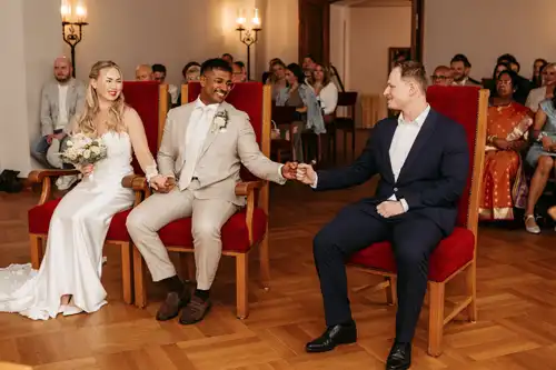 Braut und Bräutigam sitzen auf roten Stühlen, halten sich an den Händen und lächeln bei ihrer Hochzeit in Görlitz. Der Bräutigam gibt einem neben ihm sitzenden Mann in einem marineblauen Anzug einen Faustschlag, während die Hochzeitsgäste im Hintergrund zusehen.