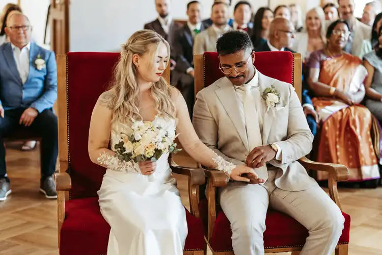 Braut und Bräutigam sitzen bei ihrer Hochzeit in Görlitz Seite an Seite auf roten Stühlen und halten sich während der Zeremonie liebevoll an den Händen, umgeben von Gästen in formeller Kleidung. Die Braut hält einen Blumenstrauß und trägt Weiß, der Bräutigam trägt einen hellen Anzug.