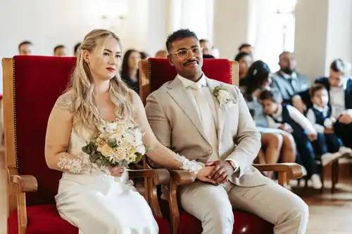 Eine Braut und ein Bräutigam sitzen auf roten Stühlen nebeneinander und halten sich bei ihrer Hochzeit in Görlitz an den Händen. Die Braut trägt ein weißes Kleid mit einem Blumenstrauß, der Bräutigam einen hellen Anzug und eine Ansteckblume. Die Gäste sitzen im Hintergrund. Hochzeitsfotograf Görlitz.