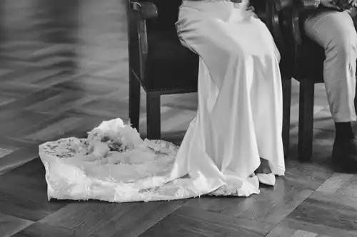 Schwarz-Weiß-Foto eines sitzenden Paares auf der Hochzeit in Görlitz, mit Fokus auf die untere Hälfte. Die Person auf der linken Seite trägt ein wallendes Brautkleid, dessen Spitze auf dem Holzboden schleift; die andere Person trägt eine helle Hose und Kleiderschuhe.