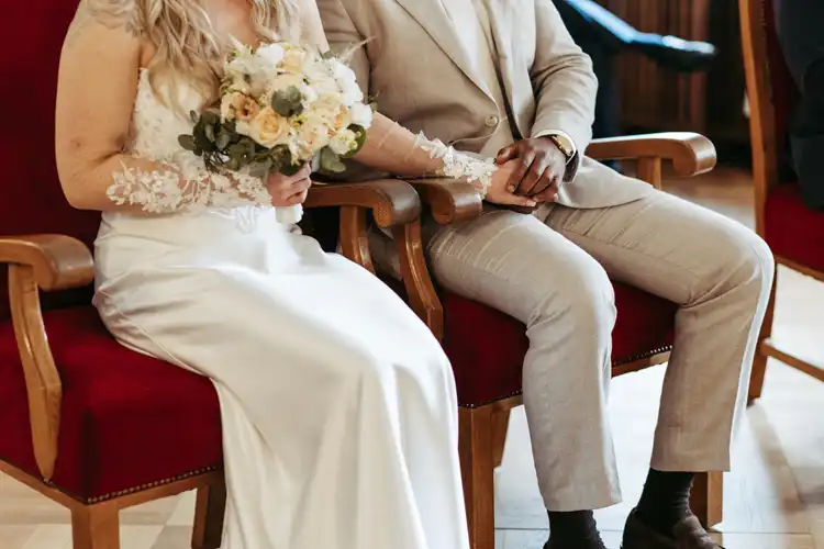 Eine Braut in einem weißen Kleid mit einem Blumenstrauß in der Hand sitzt neben einem Bräutigam in einem hellen Anzug bei ihrer Hochzeit in Görlitz. Sie halten sich auf roten Samtstühlen an den Händen, nur Torsos und Hände sind zu sehen. Aufgenommen von Hochzeitsfotograf Görlitz.