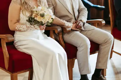 Eine Braut in einem weißen Kleid mit einem Blumenstrauß in der Hand sitzt neben einem Bräutigam in einem hellen Anzug bei ihrer Hochzeit in Görlitz. Sie halten sich auf roten Samtstühlen an den Händen, nur Torsos und Hände sind zu sehen. Aufgenommen von Hochzeitsfotograf Görlitz.