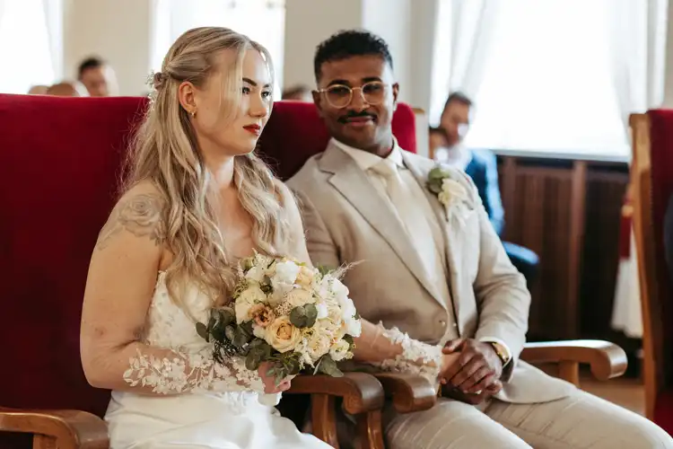 Eine Braut in einem weißen Kleid mit einem Blumenstrauß sitzt neben einem Bräutigam in einem hellen Anzug. Auf ihrer Hochzeit Gut am See sitzen sie auf roten Stühlen und halten sich während einer scheinbar herzlichen Hochzeitszeremonie an den Händen.