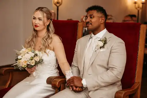 Eine Braut in einem weißen Kleid und ein Bräutigam in einem beigen Anzug sitzen nebeneinander auf roten Stühlen, halten sich an den Händen und schauen nach vorne während ihrer Hochzeit in Görlitz, wunderschön eingefangen von Hochzeitsfotograf Görlitz.