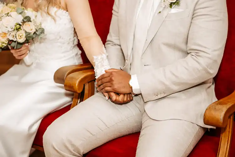 Eine Braut im weißen Kleid und ein Bräutigam im hellen Anzug sitzen auf roten Stühlen und halten sich während ihrer Hochzeit auf Gut am See an den Händen, perfekt eingefangen von Hochzeitsfotograf Görlitz.