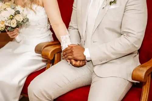 Eine Braut im weißen Kleid und ein Bräutigam im hellen Anzug sitzen auf roten Stühlen und halten sich während ihrer Hochzeit auf Gut am See an den Händen, perfekt eingefangen von Hochzeitsfotograf Görlitz.