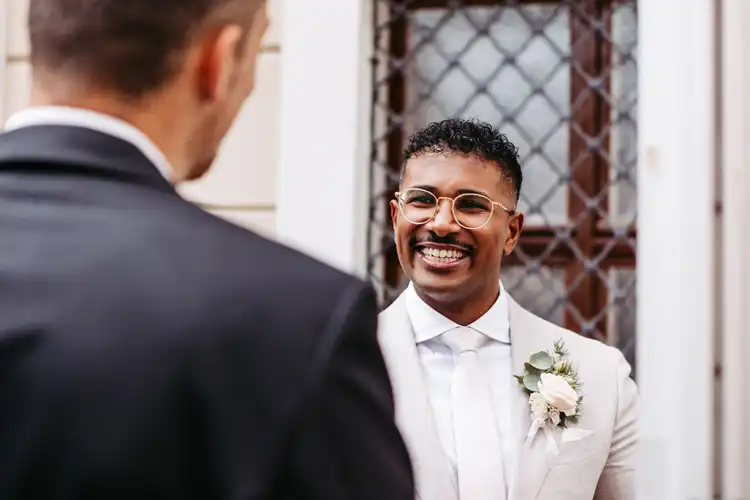 Ein Mann mit Brille, weißem Anzug und Boutonniere lächelt einer anderen Person in einem dunklen Anzug freundlich zu. Er steht im Freien auf Hochzeit Gut am See vor einem Gebäude mit einem Fenster und dekorativen Eisenbeschlägen.