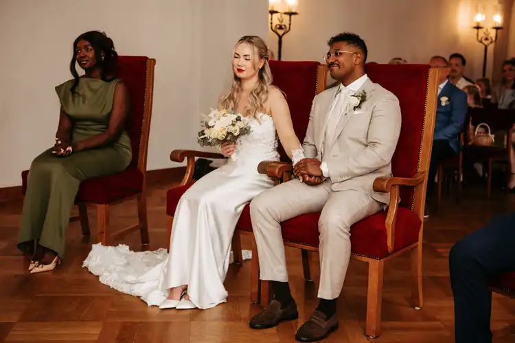 Eine Braut im weißen Kleid und ein Bräutigam im hellen Anzug sitzen während ihrer Hochzeit in Görlitz händchenhaltend auf roten Stühlen, daneben eine Brautjungfer in Grün. Der Raum ist mit Holzböden und leuchtenden Wandleuchtern ausgestattet - aufgenommen von Hochzeitsfotograf Görlitz.