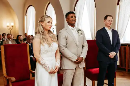 Eine Braut in einem weißen Kleid und ein Bräutigam in einem beigen Anzug lächeln sich bei ihrer Hochzeit in Görlitz an, begleitet von einem Mann in einem dunklen Anzug, in einem eleganten Raum mit großen Fenstern und roten Stühlen. Die Gäste sitzen im Hintergrund.