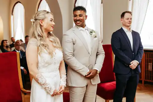 Eine Braut in einem weißen Kleid und ein Bräutigam in einem hellen Anzug lächeln gemeinsam während ihrer Hochzeit in Görlitz, mit einem Mann in einem blauen Anzug an ihrer Seite und Gästen, die dahinter sitzen - wunderschön eingefangen von Hochzeitsfotograf Görlitz.