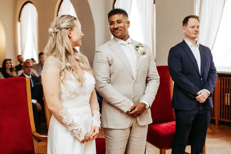 Braut und Bräutigam stehen sich lächelnd bei ihrer Hochzeit in Görlitz gegenüber, der Bräutigam in einem hellen Anzug, die Braut in einem weißen Kleid. Ein Mann in einem dunklen Anzug steht neben ihnen, während die Gäste aus dem Hintergrund zusehen.