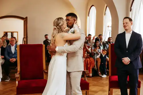 Eine Braut und ein Bräutigam umarmen sich auf ihrer Hochzeit in Görlitz, umgeben von sitzenden Gästen in einem hellen Raum. Der Bräutigam trägt einen hellen Anzug, die Braut ein weißes Kleid, und ein Mann in einem dunklen Anzug steht lächelnd daneben. Hochzeitsfotograf Görlitz fängt den Moment wunderschön ein.