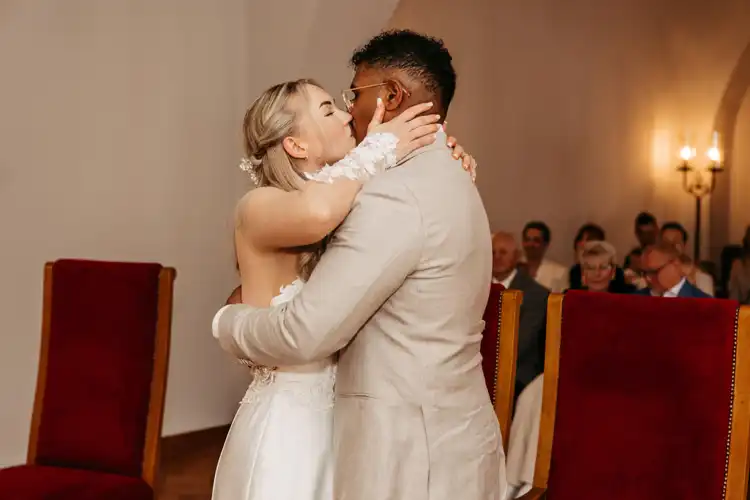 Braut und Bräutigam küssen sich während ihrer Hochzeit in Görlitz umgeben von sitzenden Gästen in einem sanft beleuchteten Raum mit roten und goldenen Stühlen.