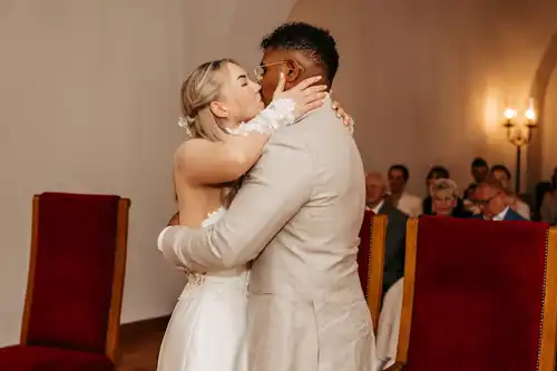 Braut und Bräutigam küssen sich während ihrer Hochzeit in Görlitz umgeben von sitzenden Gästen in einem sanft beleuchteten Raum mit roten und goldenen Stühlen.
