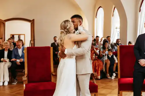 Braut und Bräutigam umarmen und küssen sich während ihrer Hochzeitszeremonie in einem sonnendurchfluteten Raum mit großen Fenstern, roten Stühlen und Gästen, die im Hintergrund sitzen.