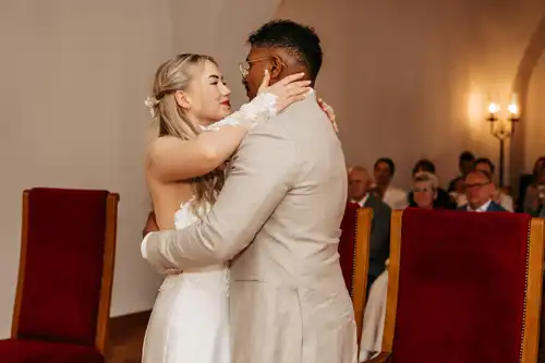 Braut und Bräutigam umarmen sich und lächeln sich während ihrer Hochzeit auf Gut am See an, während die Gäste im Hintergrund auf roten Stühlen sitzen.