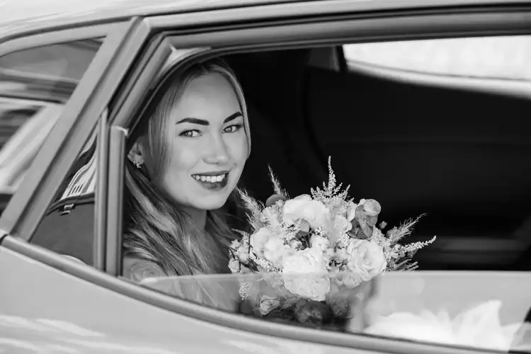 Eine lächelnde Braut, die einen Blumenstrauß in der Hand hält, sitzt auf dem Rücksitz eines Autos und blickt aus dem offenen Fenster auf ihre Hochzeit auf Gut am See. Das Schwarz-Weiß-Foto von Hochzeitsfotograf Görlitz fängt die Freude und Eleganz des Augenblicks ein.