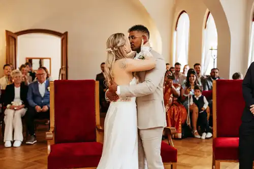 Braut und Bräutigam umarmen und küssen sich am Altar während ihrer Hochzeitszeremonie auf Gut am See in einem hellen Raum, während die Gäste im Hintergrund sitzen und zusehen.