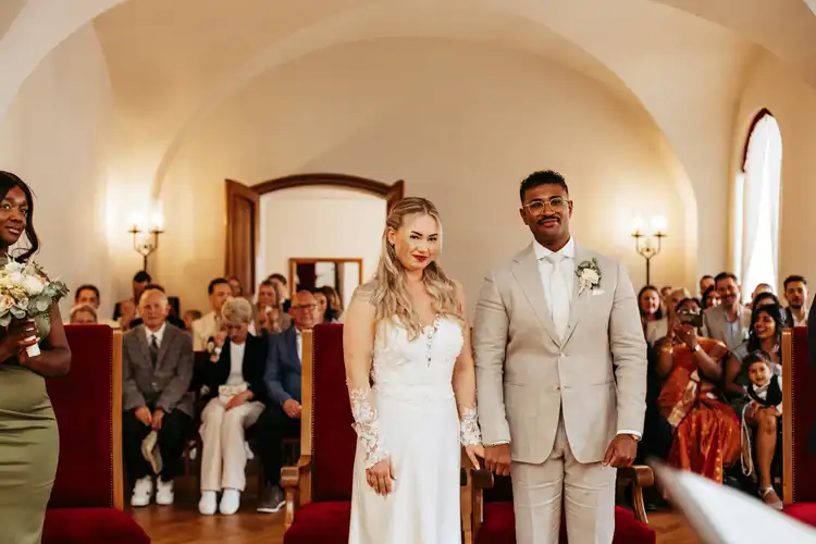 Braut und Bräutigam stehen zusammen am Altar, halten sich an den Händen und lächeln, während die Gäste in einem eleganten, gut beleuchteten Raum mit gewölbten Decken zusehen - wunderschön eingefangen von Hochzeitsfotograf Görlitz